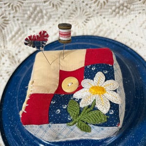 May include: A square patchwork pincushion featuring red, cream, and navy blue fabric squares with a central yellow button and an embroidered white daisy. Two pins are inserted into the top, one topped with a small heart and one with a spool.