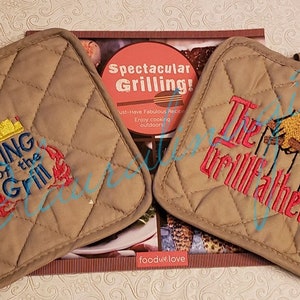 May include: Two brown quilted pot holders with embroidered designs. One pot holder says "King of the Grill" with a crown and a lobster. The other pot holder says "The Grillfather" with a hand holding a spatula and a fork.