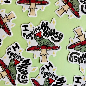 May include: A collection of stickers featuring a green frog wearing a red mushroom cap, perched on a red mushroom with white spots. The stickers have the text "I require whimsy" in a whimsical font, set against a light green background.