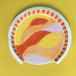 May include: A round ceramic dish with a white rim and a colorful abstract design. The design features wavy lines in shades of orange, yellow, and pink.