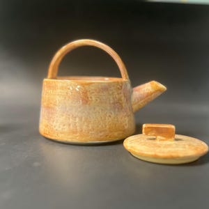 May include: A ceramic teapot with a textured, light brown glaze. The teapot has a handle and a spout. The lid is off and sitting next to the teapot.