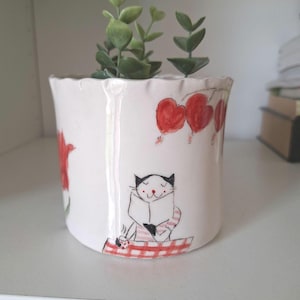 May include: White ceramic planter with a scalloped rim, decorated with red heart balloons and a cartoon cat reading a book. The planter holds a small green plant. A charming and whimsical design for home decor.