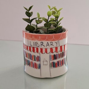 Può includere: Un piccolo vaso di ceramica bianca con un disegno di biblioteca disegnato a mano. Il vaso presenta una tenda a strisce rosse e bianche, scaffali colorati e la parola "LIBRARY". Contiene una piccola pianta verde.