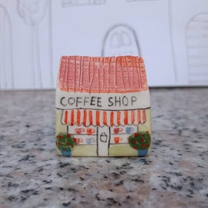 May include: A small, hand-painted ceramic coffee shop building with a red roof, white walls, and a striped awning. The shop has a sign that reads "Coffee Shop" and windows with painted coffee cups.