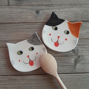 May include: Two ceramic cat-shaped spoon rests with painted faces. One cat is white with black and gray markings, the other is white with black and orange markings. Both cats have green eyes, red tongues, and pink cheeks.