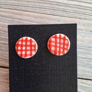 May include: Two round stud earrings with a red and white gingham pattern. The earrings are displayed on a black rectangular card against a light wood grain background. The earrings are a fun accessory.