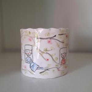 Op de afbeelding: Een witte keramische plantenpot met een gegolfde rand. De pot is versierd met een grillige illustratie van katten in ramen, omgeven door een bloemen- en takkenontwerp. De illustratie is in zachte pastelkleuren.