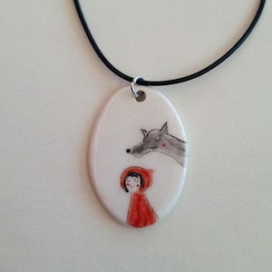 Op de afbeelding: Ovale hangerketting met een handgeschilderde illustratie van Roodkapje en een wolf. De hanger is wit met een zwart koord. Het meisje draagt een rode capuchon. De wolf is grijs.