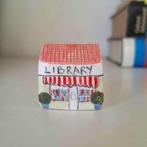 Könnte beinhalten: Miniatur-Bibliotheksfigur aus Keramik. Das Gebäude hat ein rotes Ziegeldach, eine weiße Fassade mit dem Wort "LIBRARY" über einer rot-weiß gestreiften Markise. Kleine grüne Büsche flankieren den Eingang.