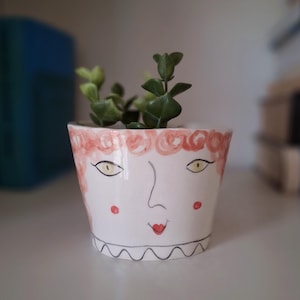 Puede incluir: Maceta de cerámica blanca con una cara pintada y pelo rojo. La maceta tiene un borde festoneado blanco y negro alrededor de la parte superior. Una pequeña planta verde está creciendo en la maceta.