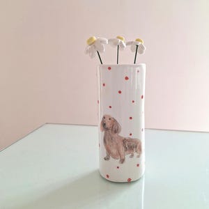 以下が含まれることがあります： 小さな赤い水玉模様と手描きの茶色のダックスフントが描かれた円筒形の白い陶器の花瓶。黄色い中心部を持つ3本の白い陶器のデイジーの花が、細い暗い色の茎に付いて花瓶の中に挿されています。