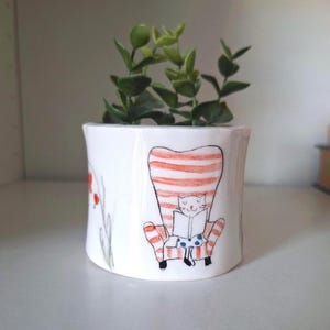 Peut inclure: Pot de fleurs en céramique blanche avec une illustration peinte à la main d'un chat lisant dans un fauteuil à rayures. Le pot présente également des motifs floraux rouges et verts et est rempli de faux feuillage vert.