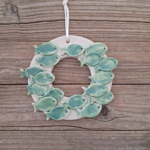 Puede incluir: Un adorno de pared circular de cerámica con una base blanca decorada con numerosas figuras de peces pequeños en color verde azulado claro dispuestas en círculo. Incluye un cordón blanco en la parte superior para colgarlo verticalmente.