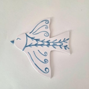 Puede incluir: Un adorno de pájaro de cerámica blanca con arte lineal azul pintado a mano. El diseño incluye un patrón de hojas estilizado a lo largo del centro del cuerpo y líneas decorativas en espiral en las alas. El pájaro tiene un pequeño pico azul y un ojo cerrado.