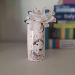 Könnte beinhalten: Weiße Keramikvase mit roten Punkten und einer handgemalten Illustration eines Husky-Hundes. Eine weiße Blume steht in der Vase.