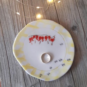 May include: A white ceramic plate with a yellow and white geometric pattern. The plate has a red flower design and a gold ring in the center. There are small black ants painted around the edge of the plate.