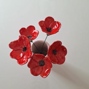 Könnte beinhalten: Draufsicht auf eine kleine, hellrosa Vase mit fünf leuchtend roten Keramik-Mohnblumen. Die Blüten haben schwarze Zentren und sind kreisförmig angeordnet, wobei die Vase im Mittelpunkt steht.