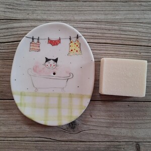 Può includere: Portasapone ovale in ceramica con un design a forma di gatto in una vasca da bagno. Il piatto presenta un'illustrazione disegnata a mano di un gatto in una vasca con bolle rosa e tre capi di abbigliamento appesi a un filo. Una saponetta è accanto al piatto.
