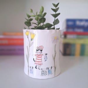 Può includere: Un vaso per piante in ceramica bianca con un'illustrazione dipinta a mano di un gatto che indossa una maglietta a righe rosse e bianche e un cappello a motivi. Il gatto è in equilibrio su un filo da bucato con panni appesi.