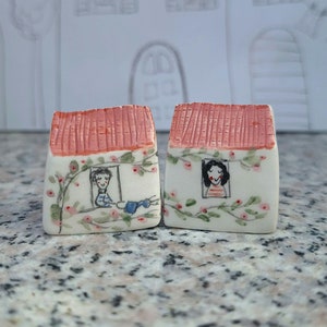 Peut inclure: Deux petites maisons en céramique avec des toits rouges et des murs blancs. Chaque maison a une fenêtre avec une personne qui regarde dehors. Les maisons sont décorées de vignes vertes et de fleurs roses.