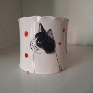 May include: White ceramic mug with a black and white cat illustration and red polka dots. The cat is depicted in profile, with a black head and white body. The mug has a wavy rim.