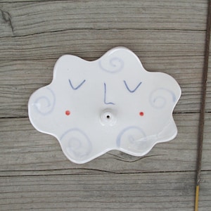 Può includere: Un porta incenso in ceramica bianca a forma di nuvola con finitura lucida. La superficie presenta un volto dipinto a mano con occhi chiusi blu, un naso semplice e due piccoli punti rossi per le guance, insieme a motivi a spirale azzurri.