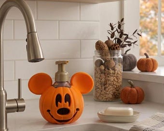 May include: An orange soap dispenser shaped like a pumpkin with round ears and a smiling face. The dispenser features a gold-colored pump mechanism on top. The pumpkin body has cutouts for the eyes, nose, and mouth, creating a festive appearance.