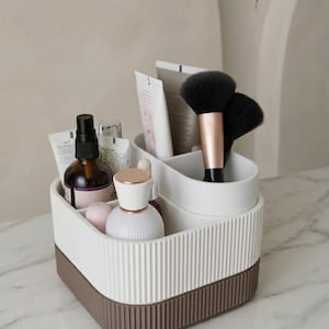 May include: A white and brown cosmetic organizer filled with beauty products, including bottles, tubes, and makeup brushes. The organizer has a ribbed design and the text "HANDCRAFTED. UNLABELED. SIMPLY CHIC."