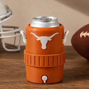 May include: An orange beverage cooler shaped like a miniature water jug, featuring a white longhorn steer logo on the front, two small white side handles, and a white spigot at the base, holding a silver aluminum beverage can inside the top.