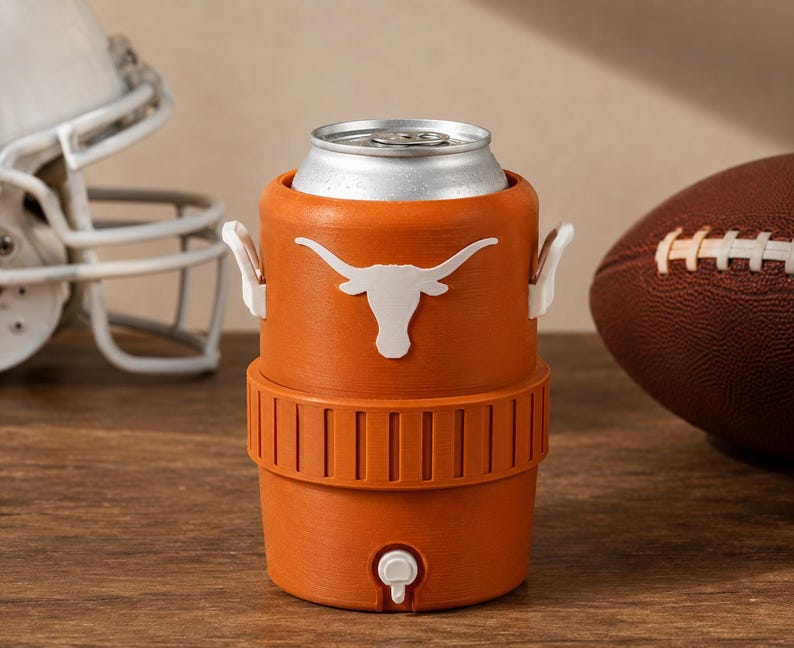 May include: An orange beverage cooler shaped like a miniature water jug, featuring a white longhorn steer logo on the front, two small white side handles, and a white spigot at the base, holding a silver aluminum beverage can inside the top.