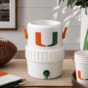 May include: A white plastic beverage cooler featuring two orange side handles and a green spigot at the base. The front of the cooler displays a large orange and green letter U logo. The cooler has a cylindrical shape with a ribbed middle section.