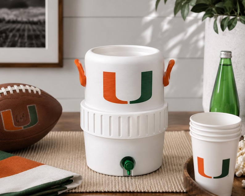 May include: A white plastic beverage cooler featuring two orange side handles and a green spigot at the base. The front of the cooler displays a large orange and green letter U logo. The cooler has a cylindrical shape with a ribbed middle section.