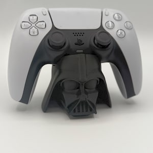 May include: A white and black PlayStation 5 controller resting on a black Darth Vader-themed stand. The controller has a symmetrical design with buttons and joysticks. The stand is a detailed replica of Darth Vader's helmet.
