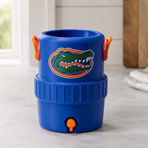 May include: A blue cylindrical beverage cooler featuring a graphic of a green alligator head with orange accents. The cooler has two orange side handles, a ribbed middle section, and an orange spigot located at the bottom front of the container.