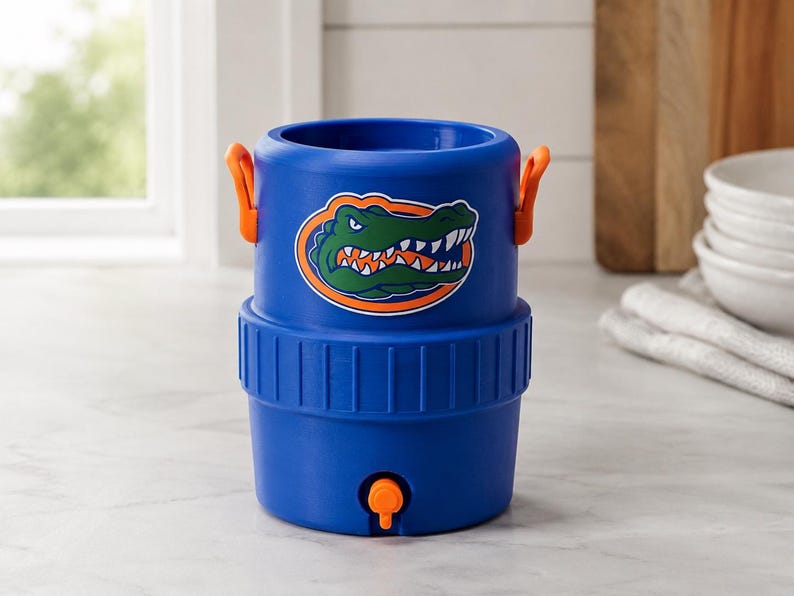 May include: A blue cylindrical beverage cooler featuring a graphic of a green alligator head with orange accents. The cooler has two orange side handles, a ribbed middle section, and an orange spigot located at the bottom front of the container.