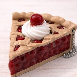May include: A slice of cherry pie charm with a whipped cream topping and a single cherry. The pie crust is a light brown color, and the filling is a glossy red. A silver chain is attached to the pie.