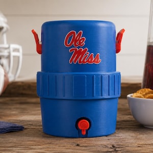 May include: A blue plastic beverage cooler featuring the red and white text Ole Miss on the front. The cooler has a red spigot at the base, two red side handles, and a textured middle band. It is a cylindrical container designed for holding liquids.