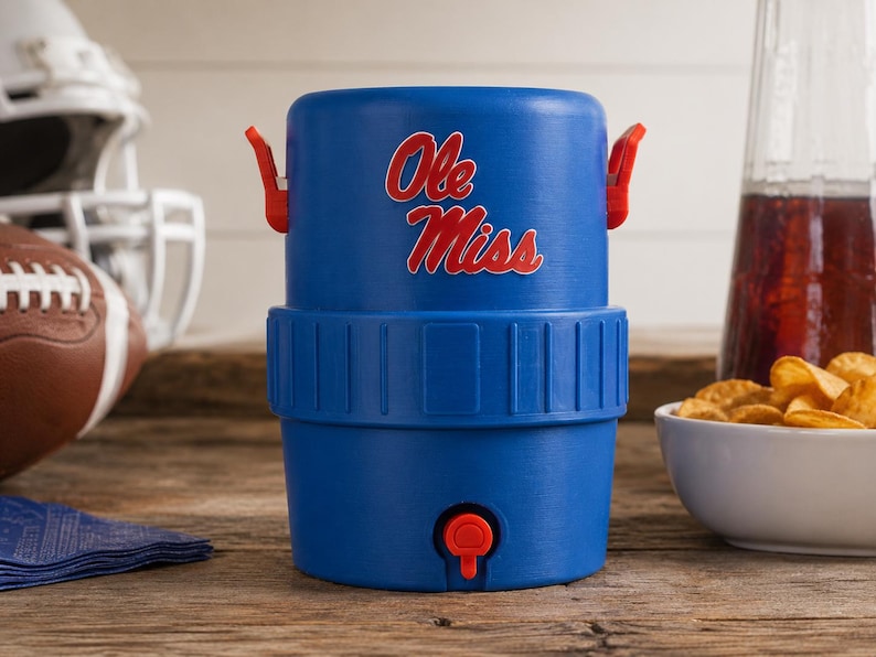 May include: A blue plastic beverage cooler featuring the red and white text Ole Miss on the front. The cooler has a red spigot at the base, two red side handles, and a textured middle band. It is a cylindrical container designed for holding liquids.