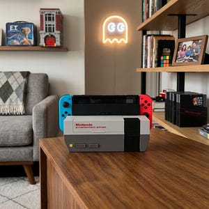 May include: A Nintendo Entertainment System-themed case with a Nintendo Switch console. The case is gray, white, and black, with the words "Nintendo Entertainment System" printed on the front. The Switch has blue and red Joy-Con controllers.