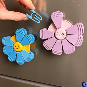 May include: Two flower-shaped magnets in blue and purple, each with a smiling face in the center. A small blue clip is held above the magnets. The magnets are attached to a gray surface, likely a refrigerator or appliance.