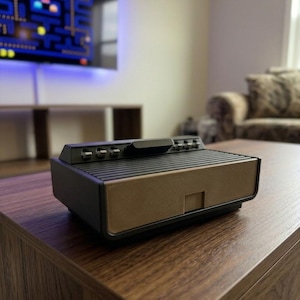 May include: A vintage video game console with a black and gold color scheme sits on a dark wood coffee table. The console has several buttons and switches. A television in the background displays a classic arcade game.