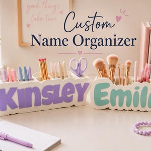 May include: Two white desk organisers with scalloped edges, each featuring a custom name in large, bold letters. The left organiser displays the name KINSLEY in purple, while the right organiser displays the name EMILIA in light teal, both filled with pens, scissors, and makeup brushes.