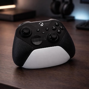 May include: A black and white Xbox controller with textured grips and buttons. The controller has a sleek design with a white bottom section and is resting on a wooden surface. A pair of black headphones are visible in the background.