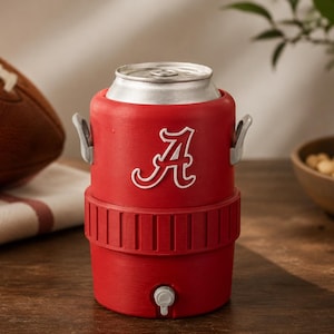 May include: A red beverage cooler shaped like a miniature water jug, featuring a white letter A logo on the front. The cooler has two small grey side handles, a textured middle band, and a small grey spigot at the base, holding a silver metal can.