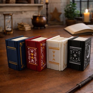 May include: Four decorative book-shaped boxes in blue, red, cream, and black, each with gold or white accents. The books have intricate designs, including moons, stars, and floral patterns. A wand lies on the wooden surface.