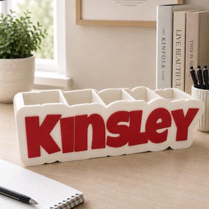 May include: A white rectangular desk organiser featuring five vertical compartments and the name KINSLEY in bold, raised red lettering on the front. The organiser has a smooth, matte finish and a scalloped edge design along the top and bottom borders.
