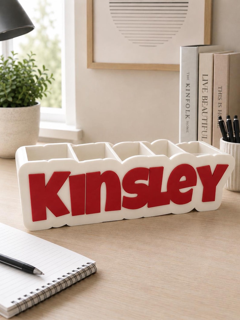 May include: A white rectangular desk organiser featuring five vertical compartments and the name KINSLEY in bold, raised red lettering on the front. The organiser has a smooth, matte finish and a scalloped edge design along the top and bottom borders.