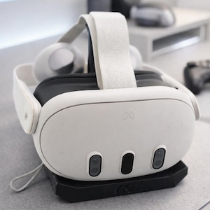 May include: A white VR headset with a black base. The headset has a white strap and ear cups. The front of the headset has three black sensors and the Meta logo. The headset is sitting on a gray surface.