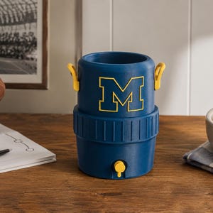 May include: A blue cylindrical beverage cooler featuring a yellow block letter M on the front. The cooler has two small yellow side handles and a yellow spigot at the base. The design includes a ribbed texture around the middle section of the container.