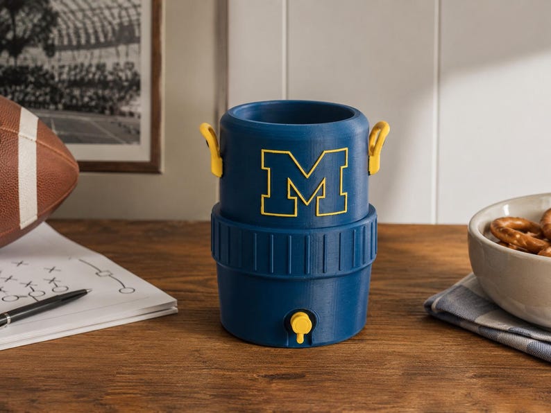 May include: A blue cylindrical beverage cooler featuring a yellow block letter M on the front. The cooler has two small yellow side handles and a yellow spigot at the base. The design includes a ribbed texture around the middle section of the container.